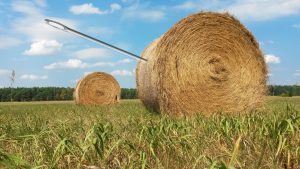 A giant needle pierces one of two haystacks on a mowed field under partly cloudy skies