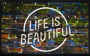 A neon sign against a brick wall. The sign reads "LIFE IS BEAUTIFUL" in all capital letters.