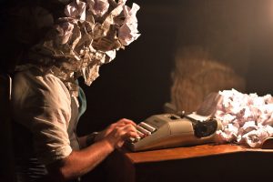 A person types on a typewriter surrounded by wadded up discarded drafts, some of which cover the typist’s head.