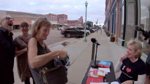 A smiling Ellen Jovin at the Grammar Table on a sidewalk in Red Cloud, Nebraska, August 2019, with three people looking on and laughing