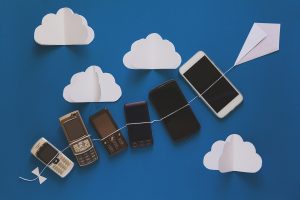 Six increasingly recent generic mobile phones, from older styles with physical buttons to newer styles resembling iPhones, all connected by a paper kite string and surrounded by paper clouds on a blue background.