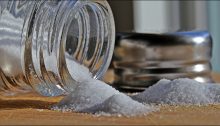 Closeup of a glass saltshaker lying on its side next to its metal screw-top lid, salt spilling out onto the table.