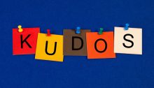 The five letters of the word “KUDOS” tacked onto a blue background on five sticky notes of various colors.