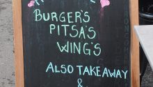 A chalkboard sidewalk sign for a restaurant in Helsinki, Finland. The sign reads "Homemade Burger's, Pitsa's, Wing's. Also Takeaway & Wolt." Note the "grocer’s apostrophe" in three places and the word "pizza" spelled as p-i-t-s-a.